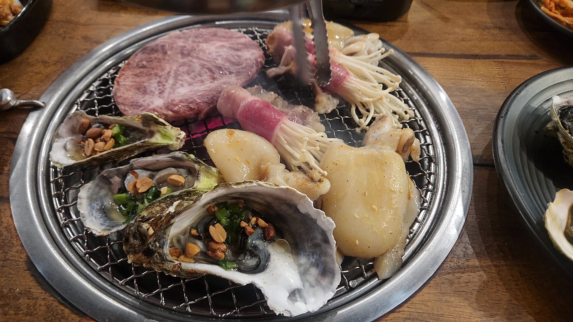 Raw beef slices, scallops, oysters, and mushrooms being grilled on a smokeless mesh grill at the table